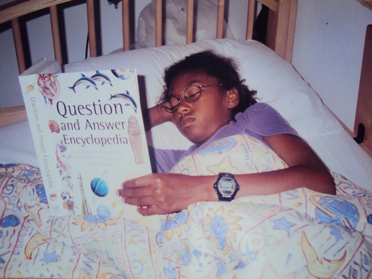 Simie as a kid in glasses, reading a Question and Answer Encyclopedia in bed
